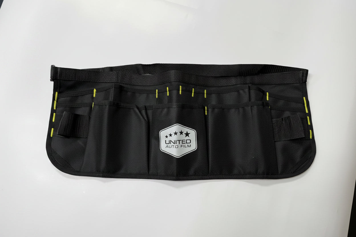 A black utility tool belt with multiple pockets, yellow stitching accents, and a white shield-shaped 'UNITED AUTO PARTS' logo patch on the front, laid flat on a white background.